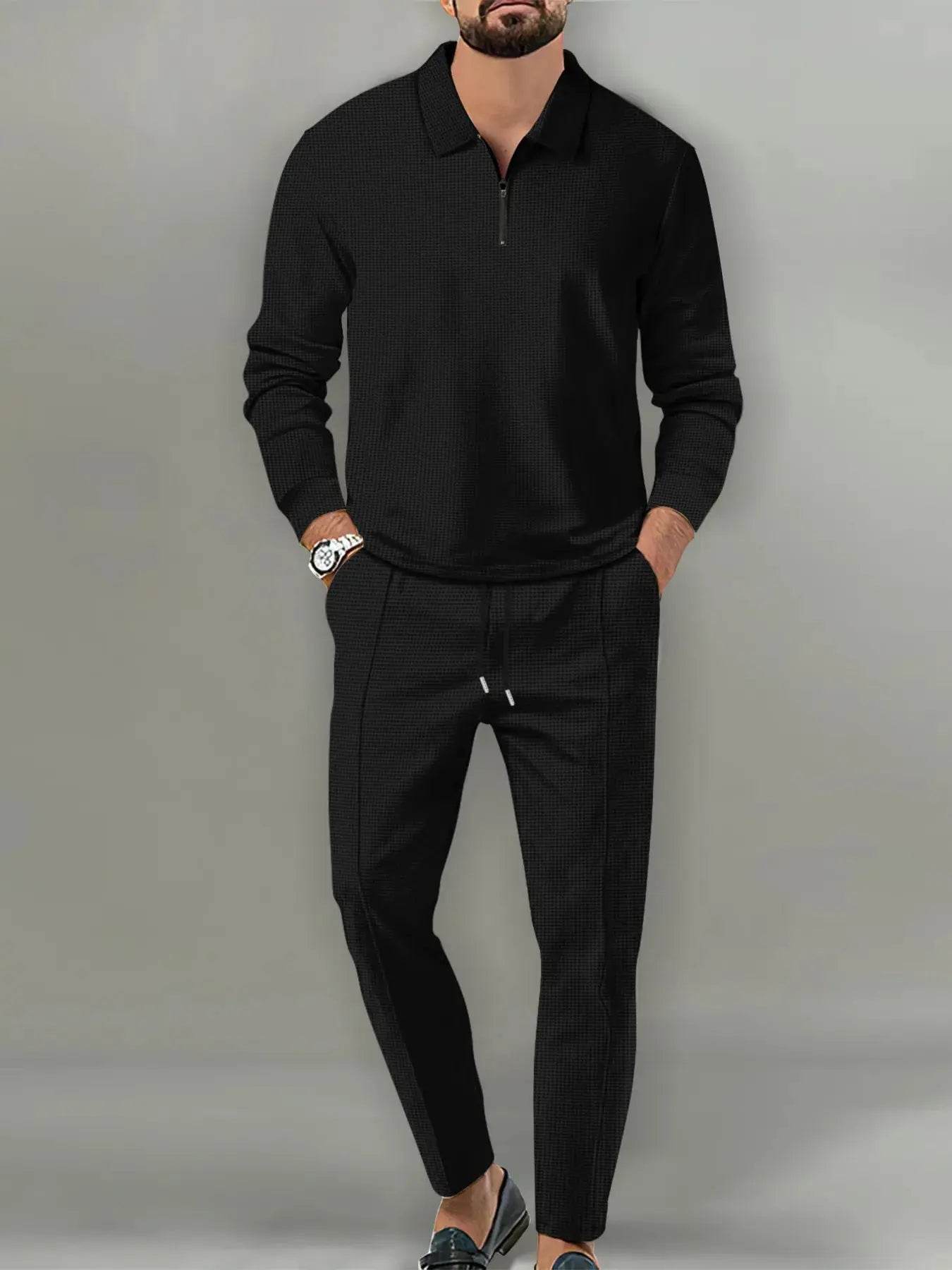 A man models a black long-sleeve collared tracksuit with a quarter-zip top and drawstring pants.