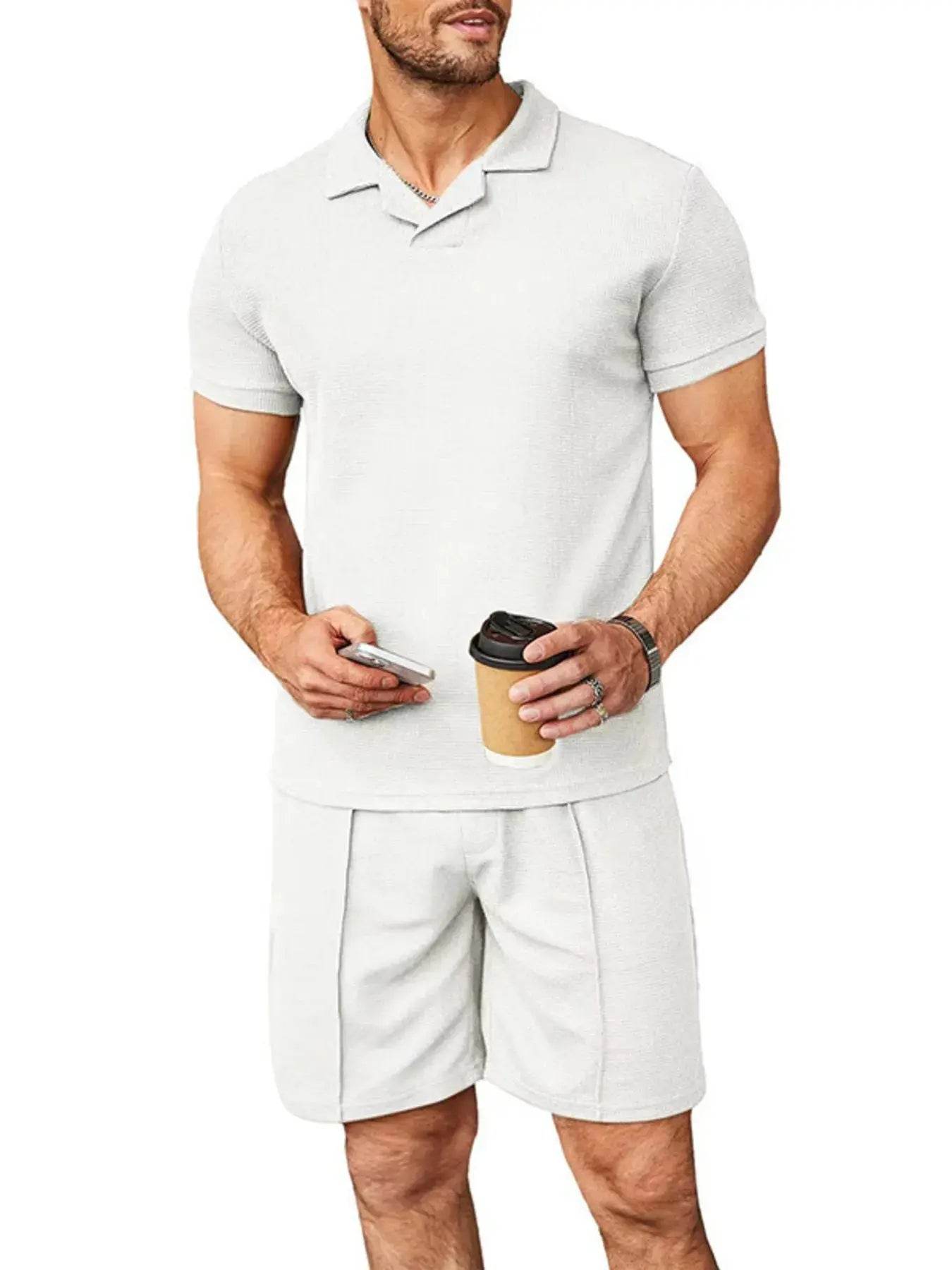 A man wears a white short-sleeve top and matching shorts, holding a phone and coffee.