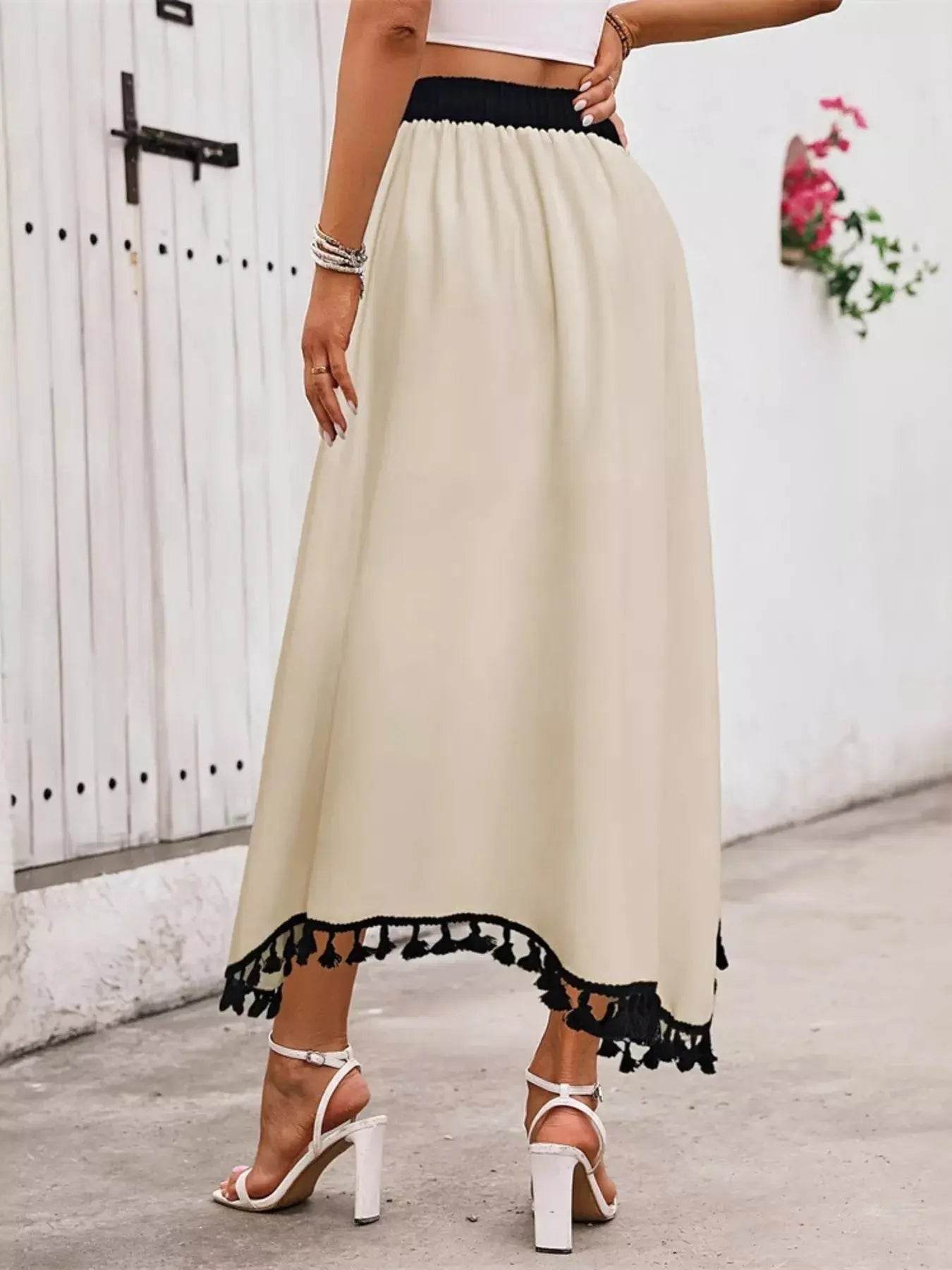 A woman models a high-waisted, beige skirt with black tassel trim and white heels.