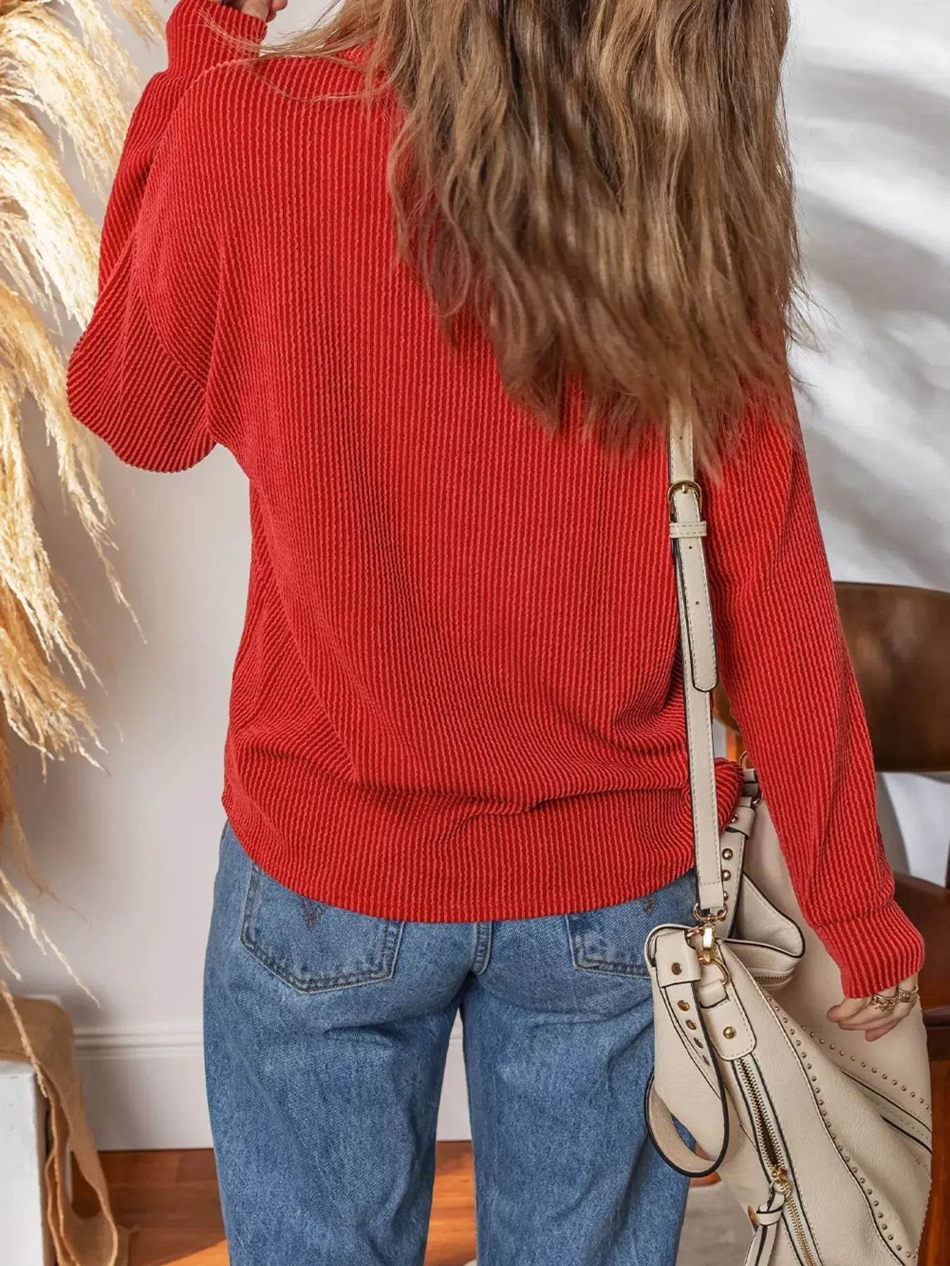 A woman from behind wears a red ribbed top, blue jeans, and carries a cream handbag.