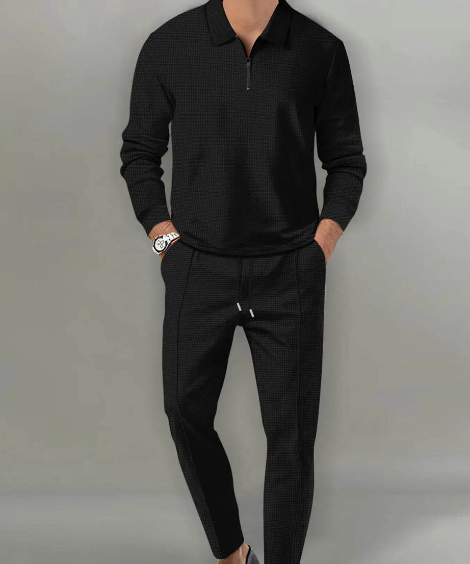 A man models a black long-sleeve collared tracksuit with a quarter-zip top and drawstring pants.