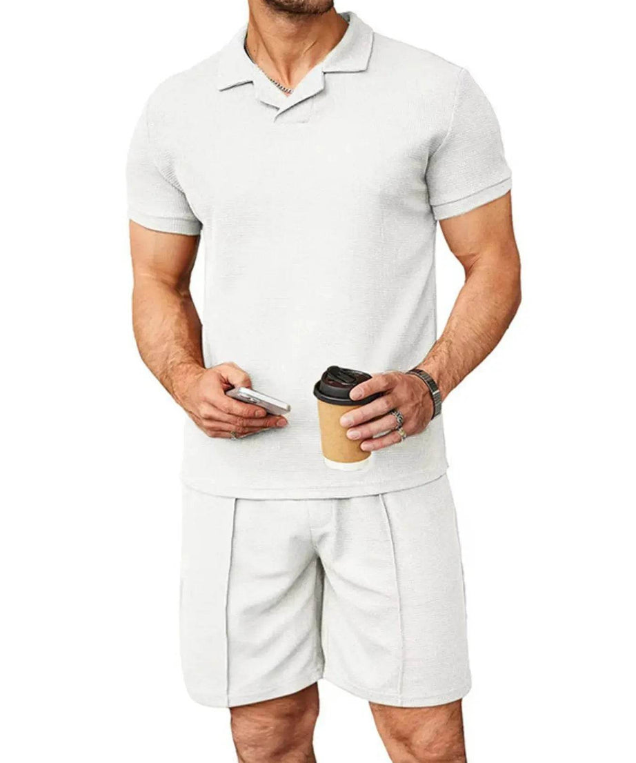 A man wears a white short-sleeve top and matching shorts, holding a phone and coffee.